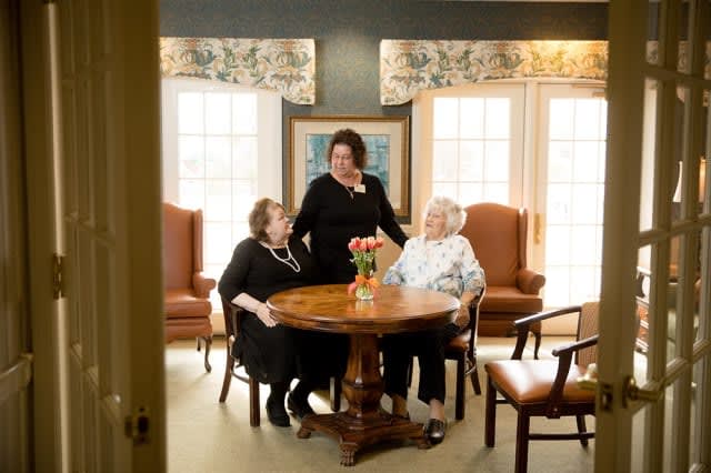Two elderly women sitting at a table with a caregiver in a care home setting.