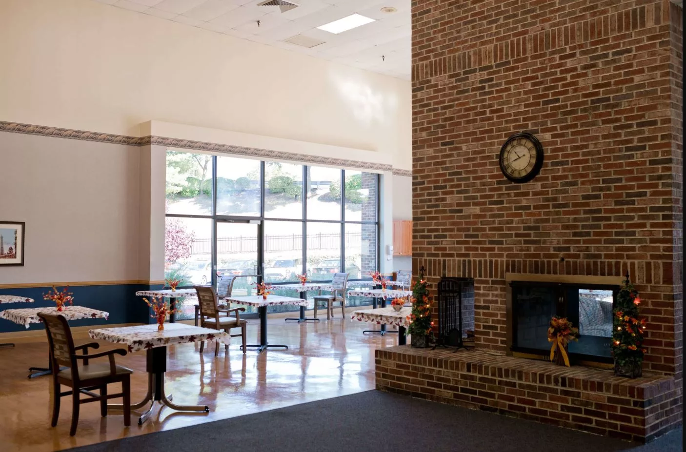 A spacious cafeteria with tables and chairs, featuring a brick fireplace and large windows.