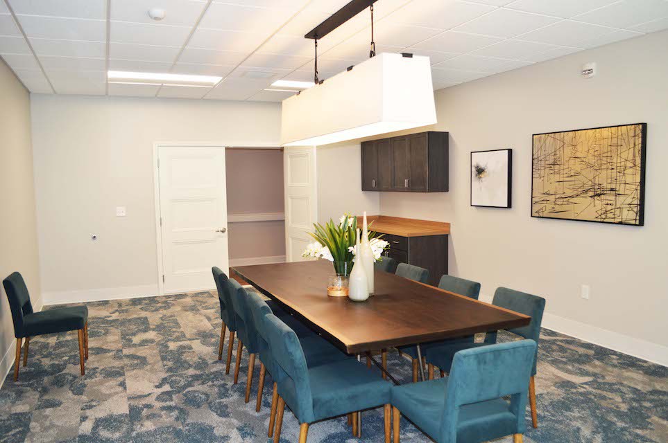 A modern meeting room featuring a long conference table surrounded by teal chairs, illuminated by a stylish overhead light fixture. The room includes wall-mounted cabinets and decorative artwork.