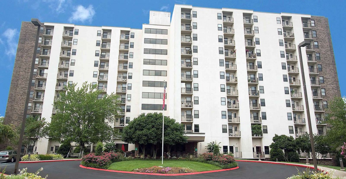 A high-rise residential apartment building with multiple balconies, surrounded by greenery.