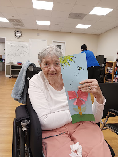 An elderly woman proudly displaying her colorful craft creation featuring a flower.