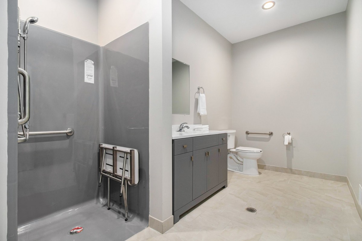 An accessible bathroom featuring gray tiles, a sink, toilet, grab bars, and a folding seat in the shower area.