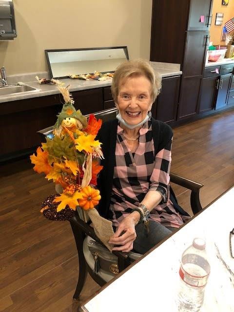 An elderly woman sitting indoors, smiling and holding autumn-themed decorations.
