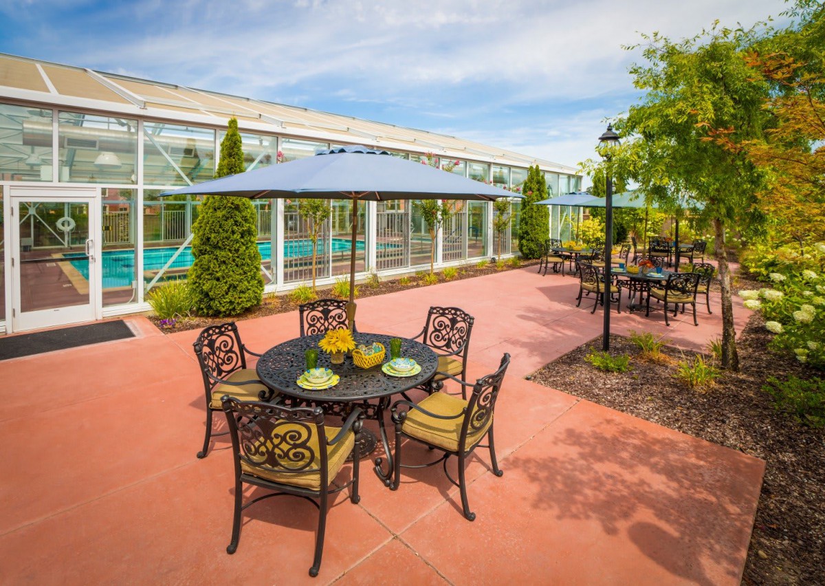 A sunny outdoor patio with tables, chairs, and an umbrella, surrounded by greenery.