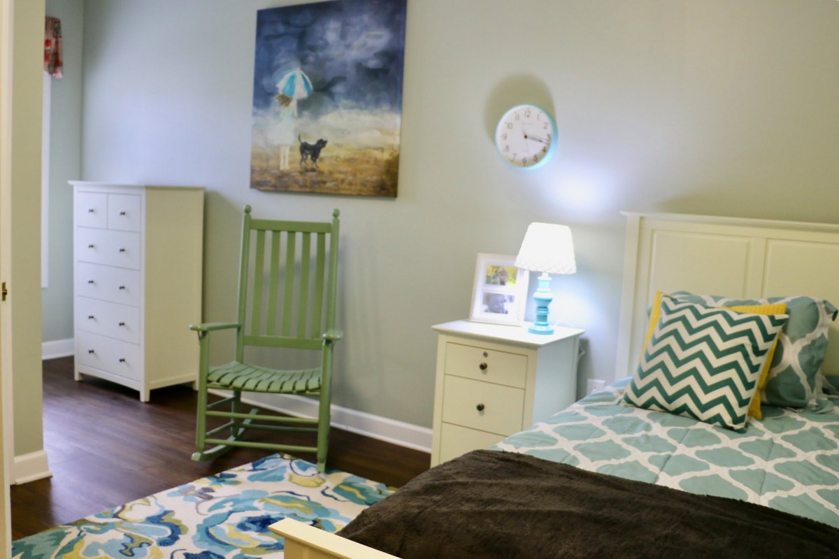 A cozy bedroom featuring a green rocking chair, a nightstand with a lamp, and artwork on the wall.