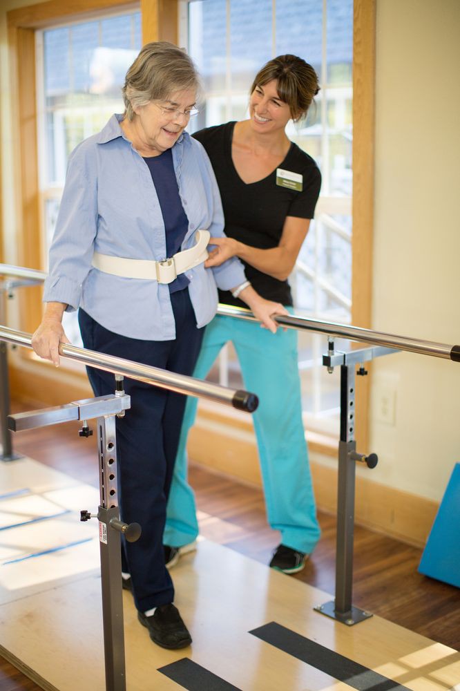 A healthcare professional assists an elderly person with physical therapy using parallel bars.