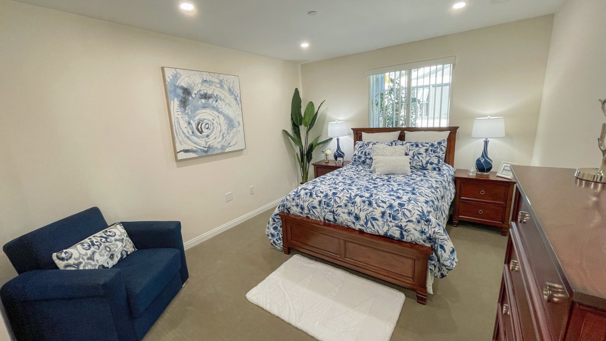 A cozy bedroom with blue floral bedding, a wooden bed frame, and a large potted plant near the window.
