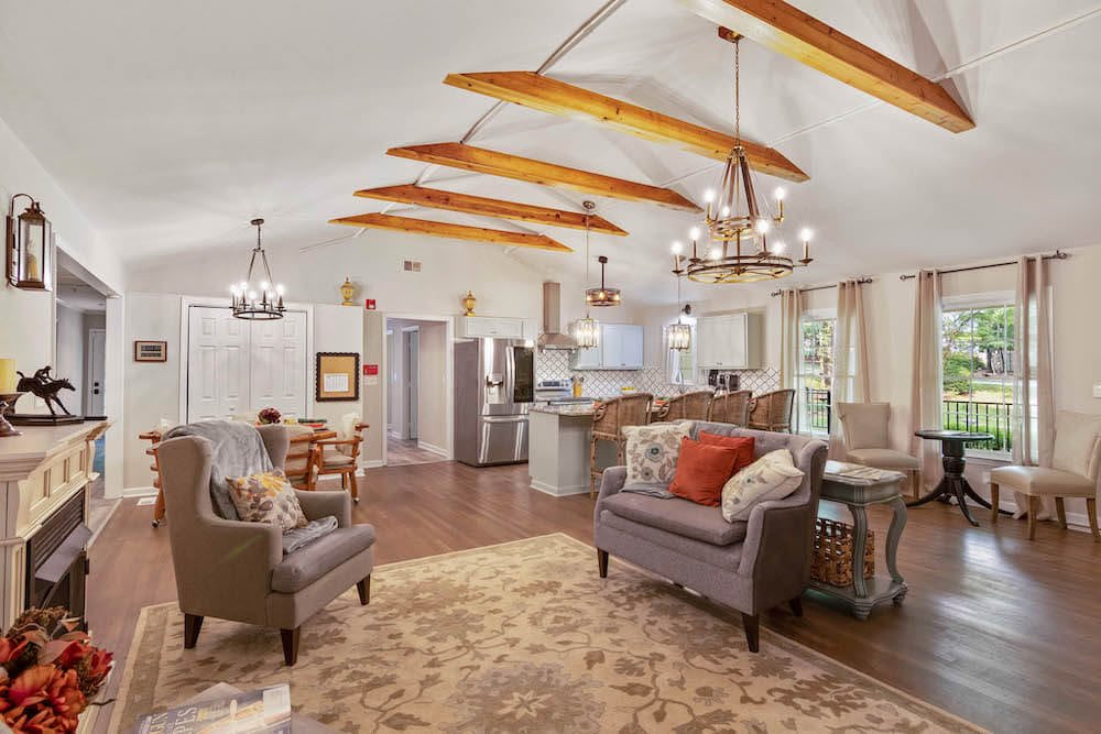 A cozy open-concept living area with vaulted ceilings and wood beams, featuring a kitchen, comfortable seating, and elegant chandeliers.