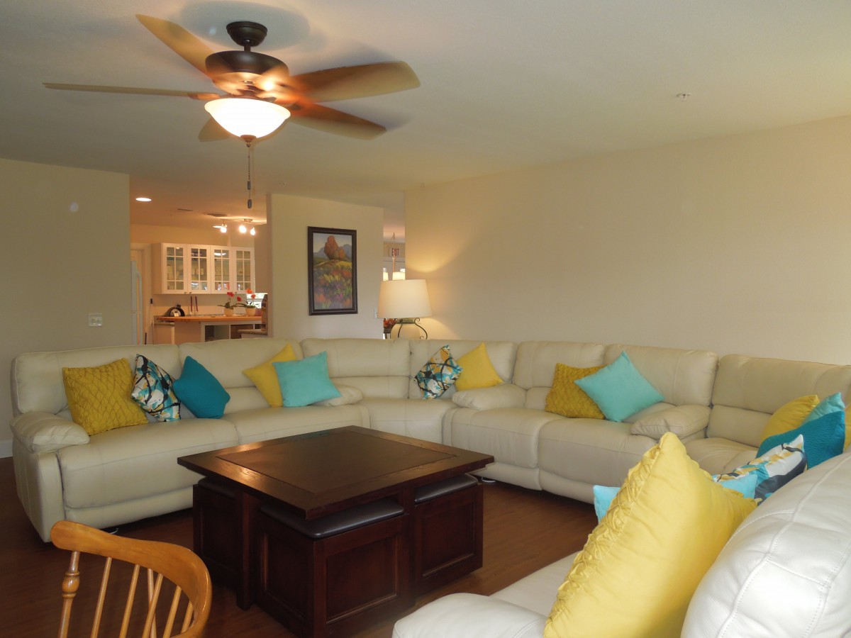 A cozy living room with a large sectional sofa adorned with colorful pillows, a central coffee table, and a ceiling fan.