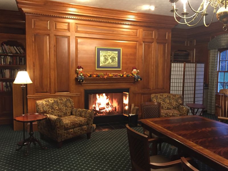 A cozy living room with wood paneling, featuring a fireplace, armchairs, and a chandelier.