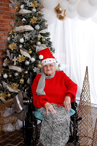 An elderly woman dressed in festive attire enjoys the holiday season by a beautifully decorated Christmas tree.
