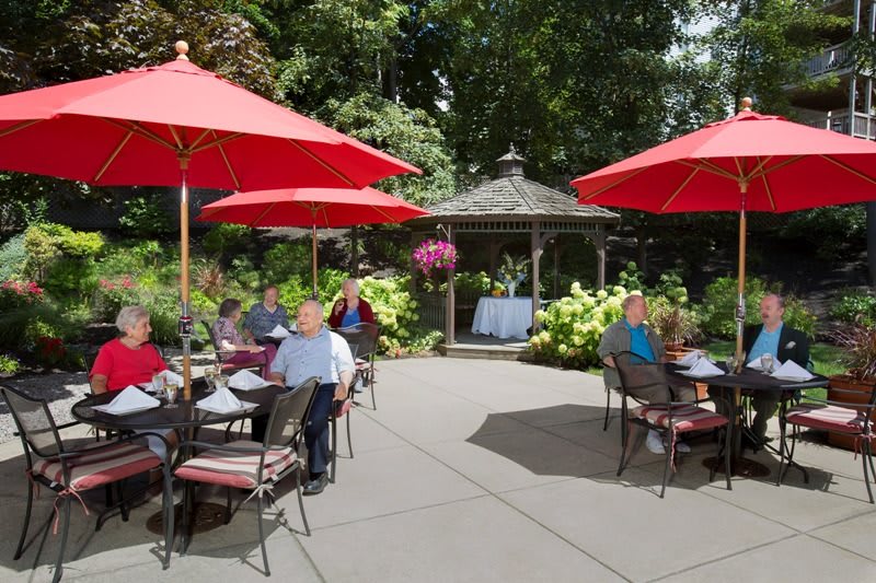 A group of people enjoying a meal on an outdoor patio with red umbrellas and a gazebo in a garden setting.