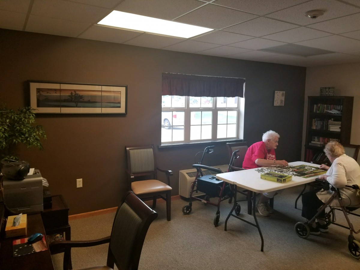 An elderly person sits at a table in a bright room, reading by the window.