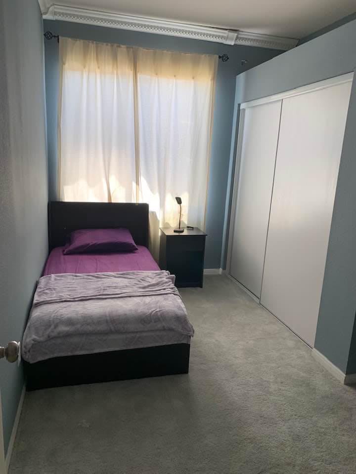 A small bedroom with a single bed, nightstand, and window with curtains.
