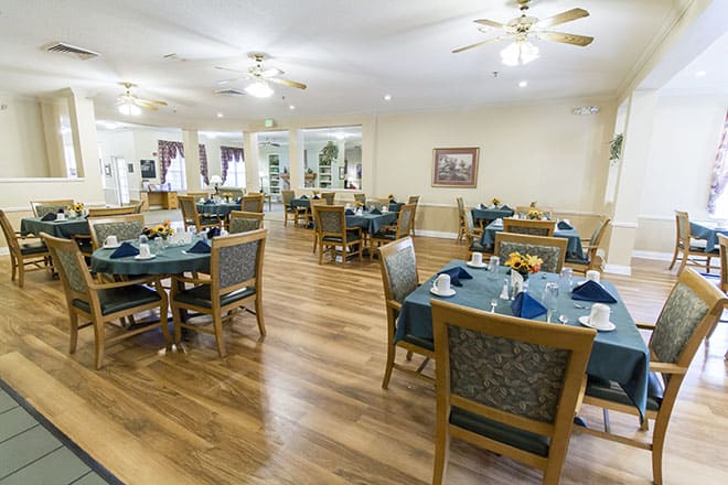 A spacious dining area with neatly arranged tables and chairs, featuring wooden floors and ceiling fans.