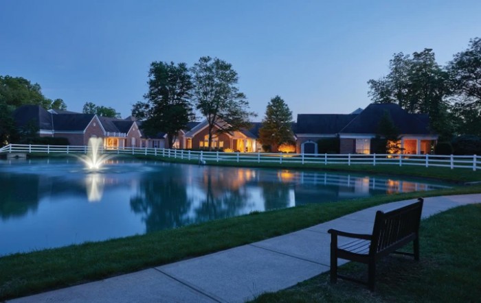 A peaceful evening scene with houses reflecting on a pond, illuminated by soft lighting and surrounded by trees and a pathway.