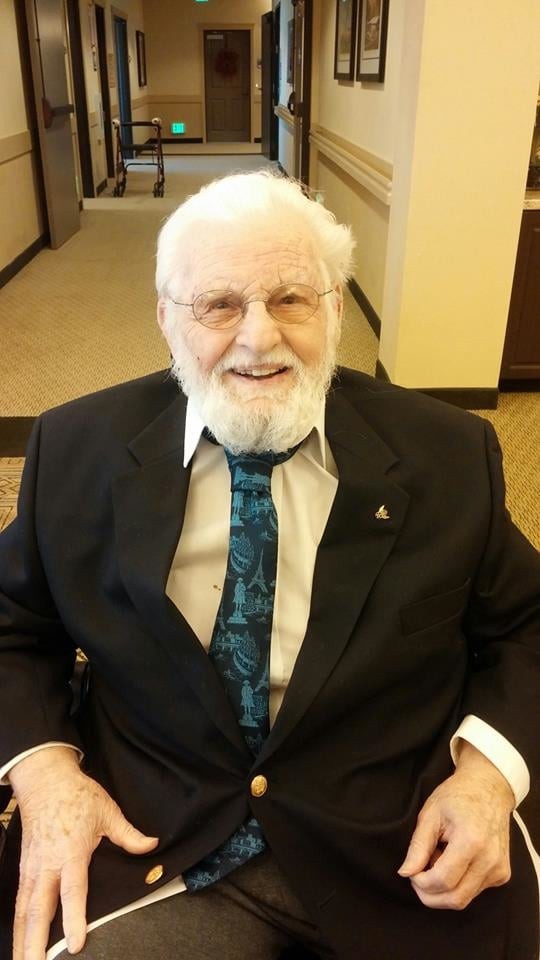 An elderly man with a beard and glasses smiling while seated indoors, wearing a suit and tie.