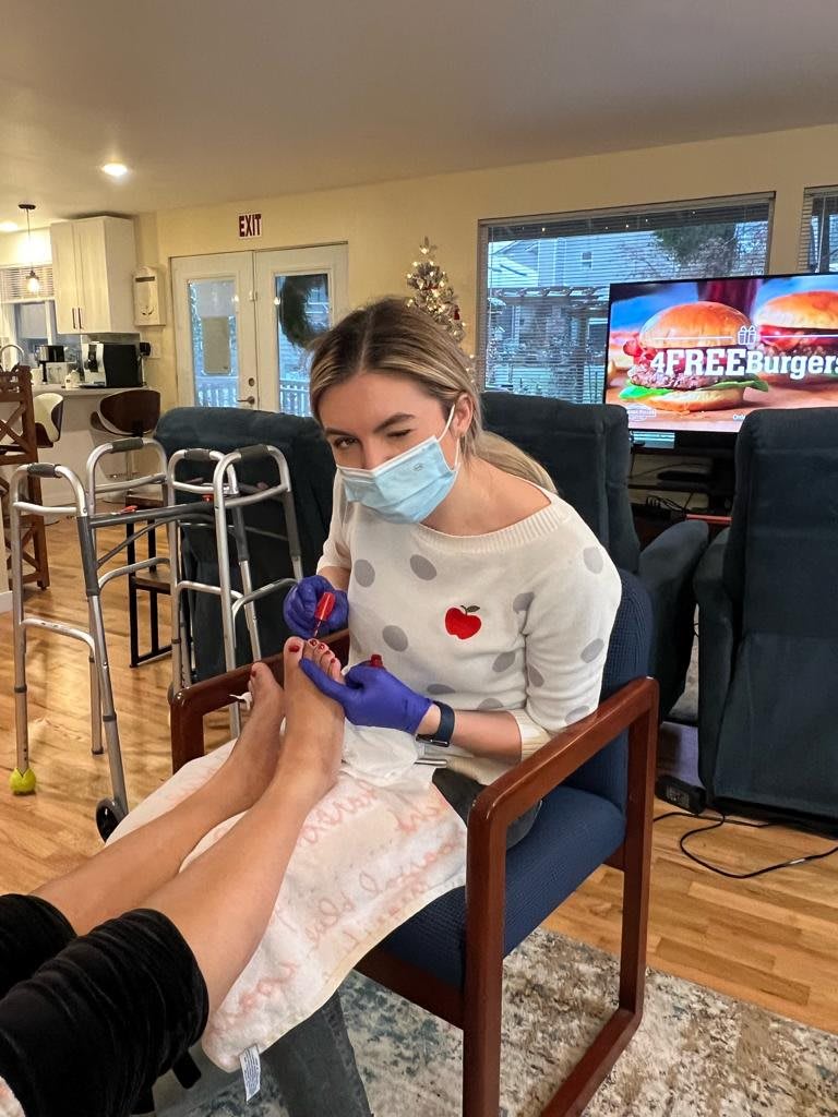 A healthcare worker provides a pedicure while wearing a mask and gloves in an indoor setting.