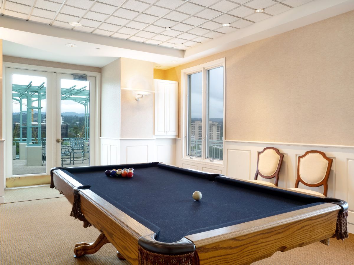 A stylish game room featuring a billiards table with a black felt cover and elegant chairs by the windows.