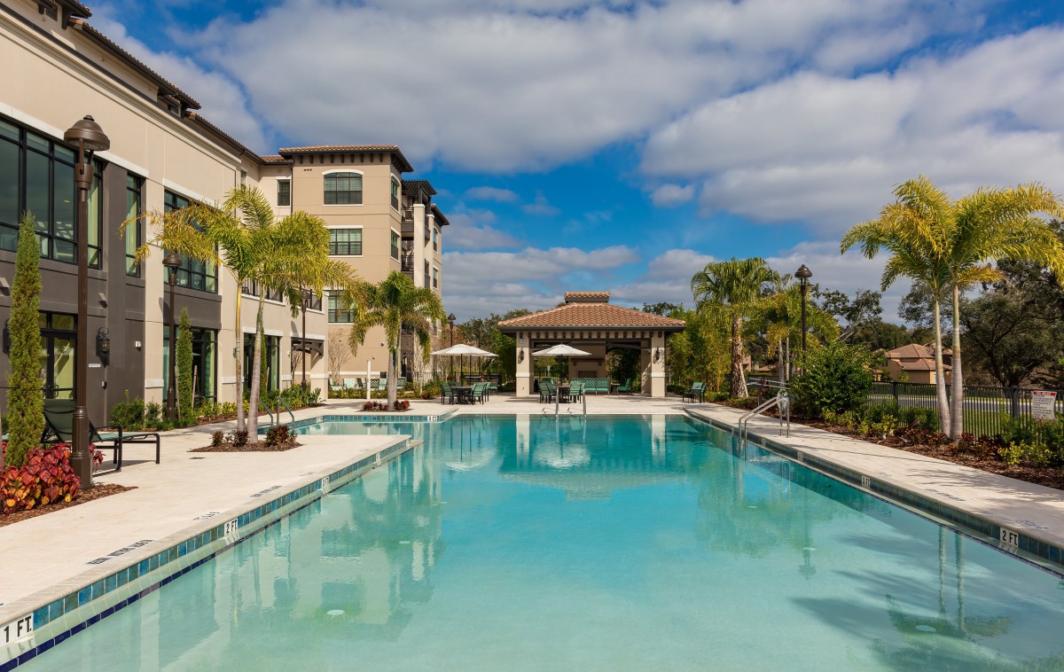 A luxurious outdoor swimming pool surrounded by a modern apartment complex and palm trees under a clear blue sky.