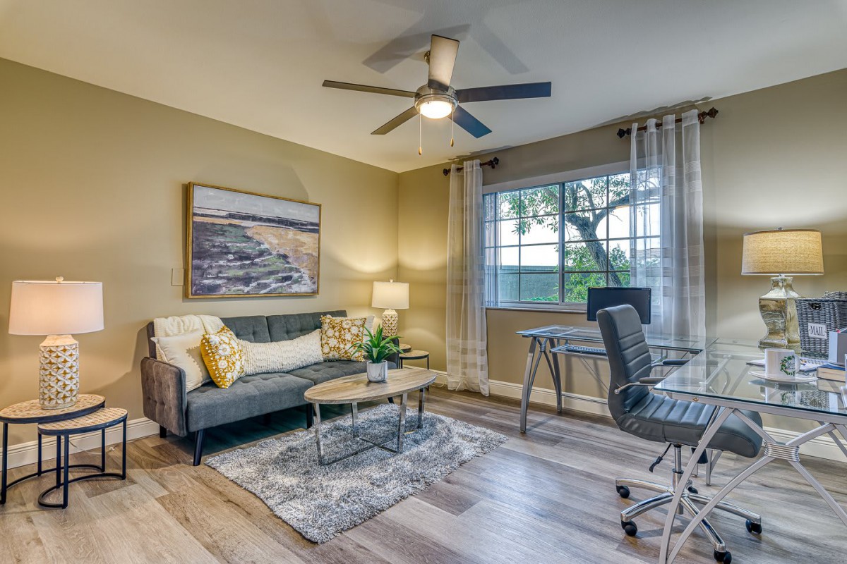 A cozy living room doubles as a home office, featuring a sofa, desk, and large window with natural light.