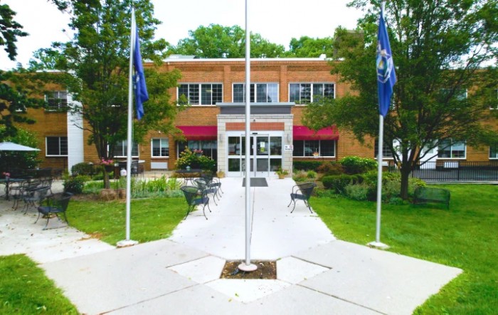 A brick building entrance with a pathway lined by flags, surrounded by trees and outdoor seating.