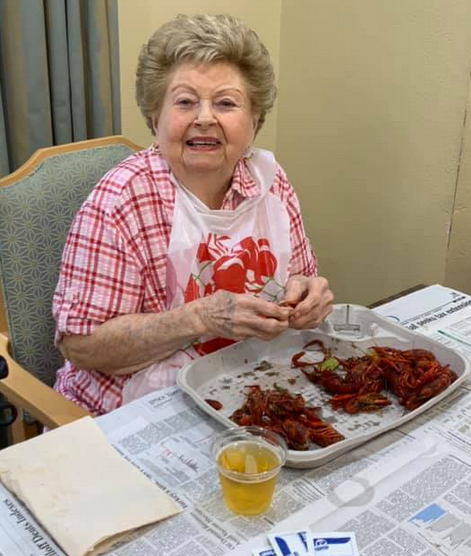 An elderly woman enjoys a meal with a smile, wearing a bib and sitting indoors with a tray of seafood.