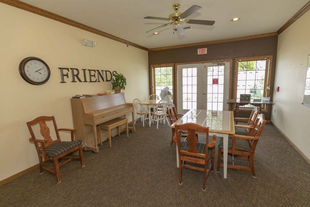 A cozy community room featuring a piano, tables, and chairs with 'Friends' decor on the wall.
