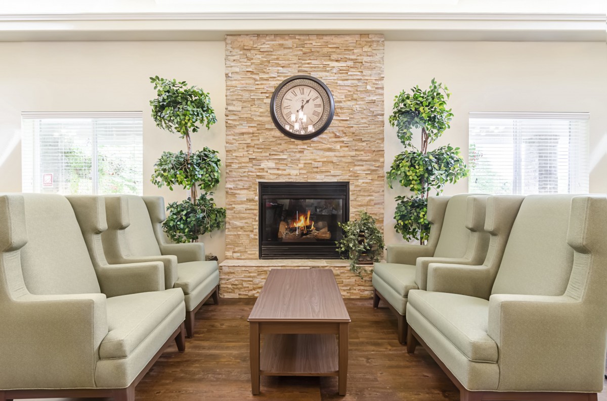 A cozy living room with a fireplace, clock, and comfortable seating surrounded by plants.