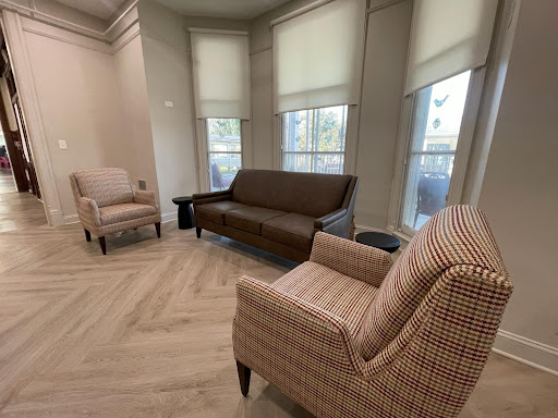 A cozy living room setup featuring a brown sofa and patterned armchairs, illuminated by natural light from large windows.