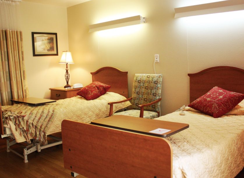 A hospital room with two beds, each equipped with a bedside table and an armchair between them.