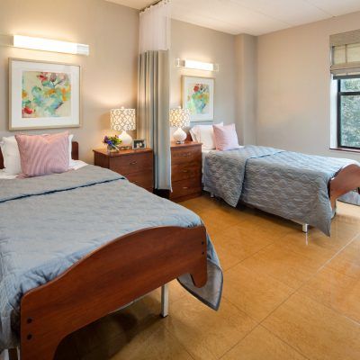 A well-organized hospital room featuring two beds with blue bedspreads, separated by a curtain and accompanied by wooden furniture and decorative elements.