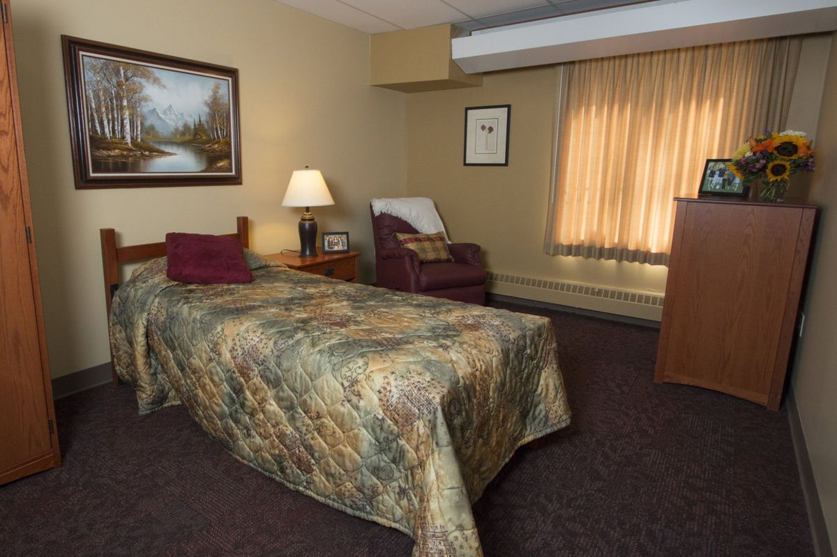 A cozy bedroom with a single bed, lamp, and armchair, featuring warm lighting and decor.