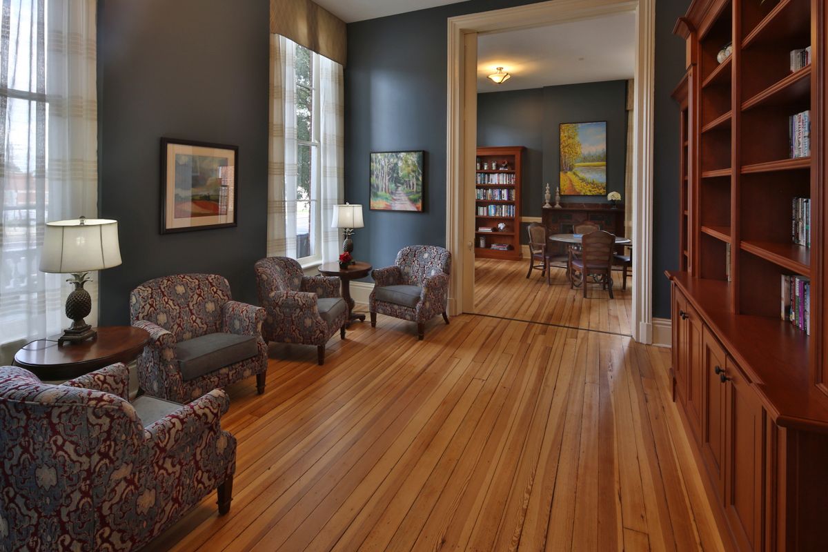 A cozy living room with armchairs, wooden floors, and large windows leading to a study area with bookshelves.