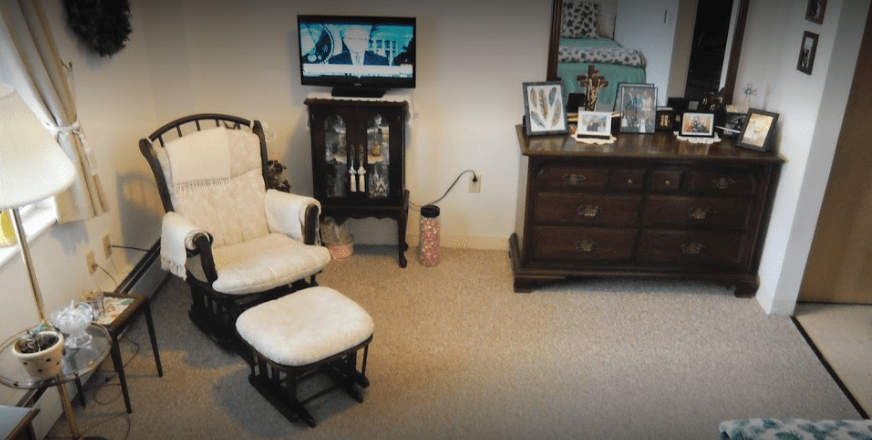 A cozy living room with a rocking chair, ottoman, TV on a stand, and a dresser displaying framed photos.