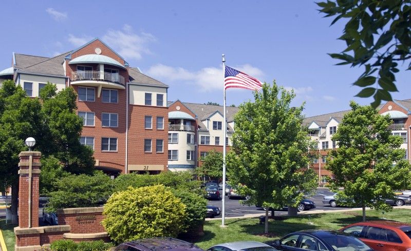 A landscaped area with a multi-story building, an American flag, and parked cars under a partly cloudy sky.