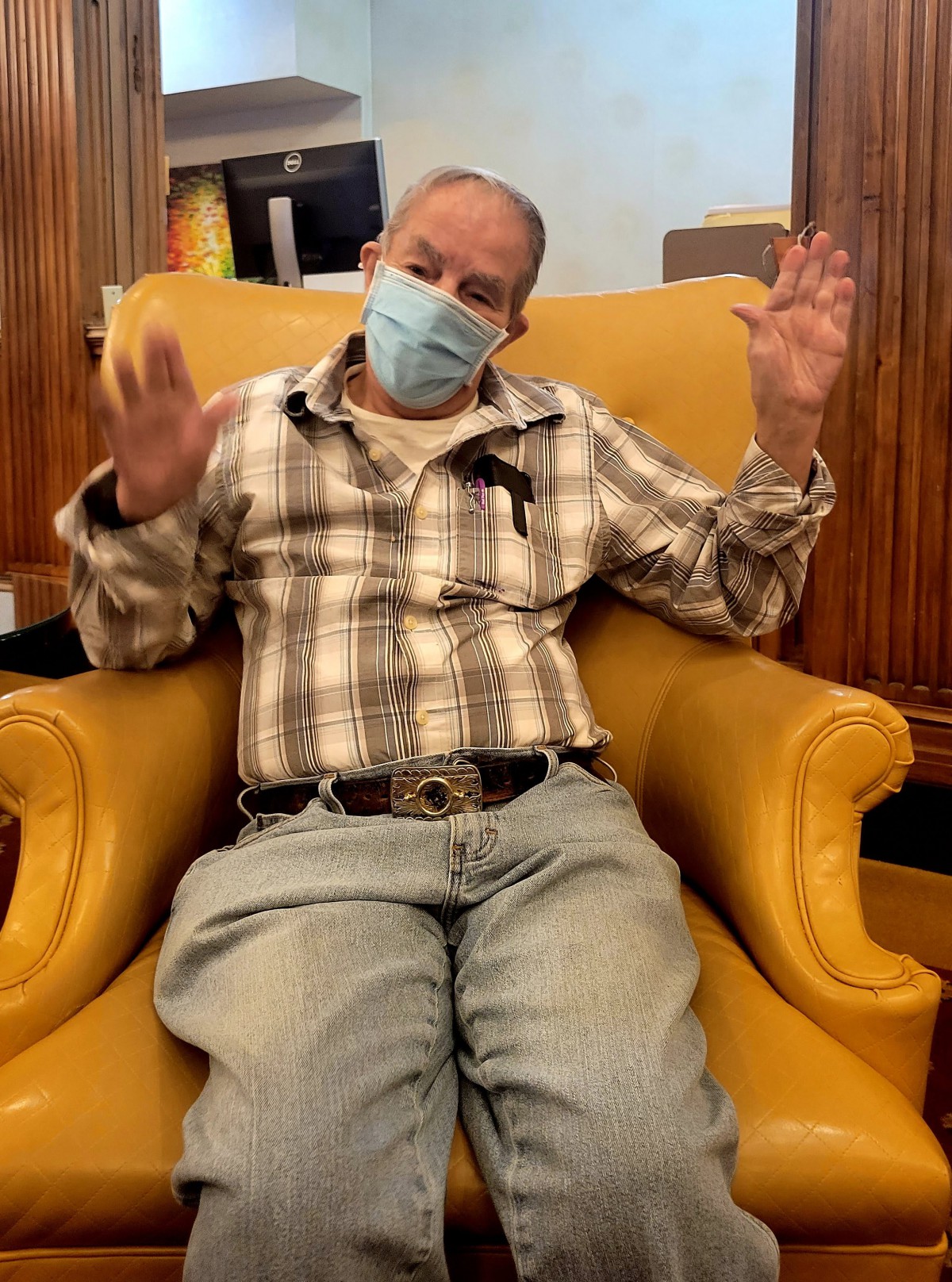 A man wearing a mask is seated in a yellow chair, wearing a plaid shirt and jeans.