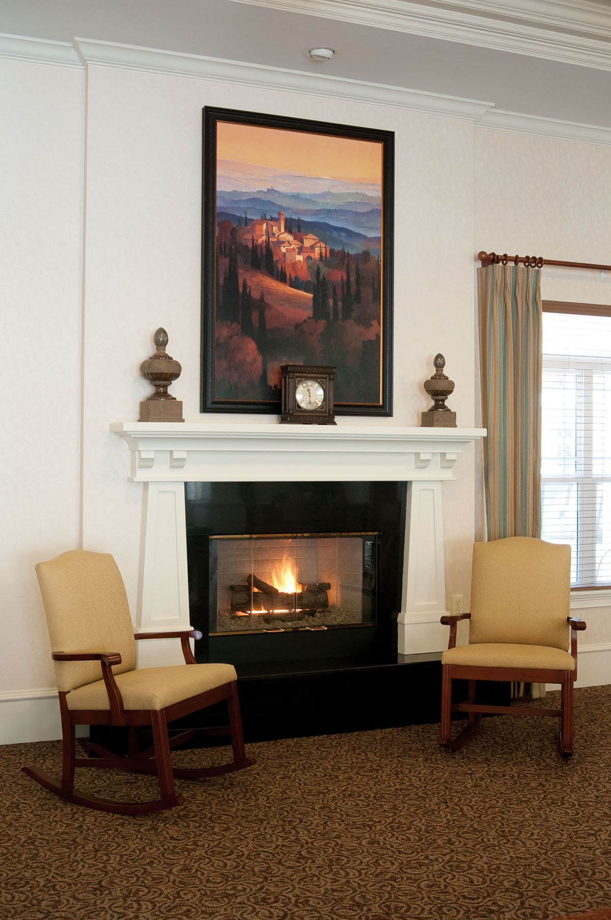 A cozy living room with a fireplace, flanked by two chairs and adorned with a landscape painting above the mantel.