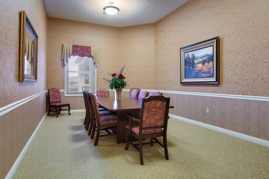 A formal dining room with a long table, surrounded by chairs, featuring a painting on the wall and a window with curtains.