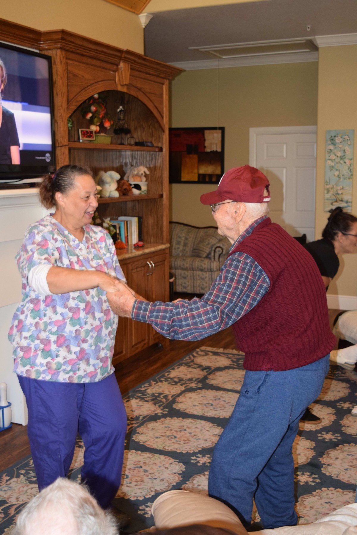 An elderly man enjoys a dance with a caregiver in a cozy living room setting.
