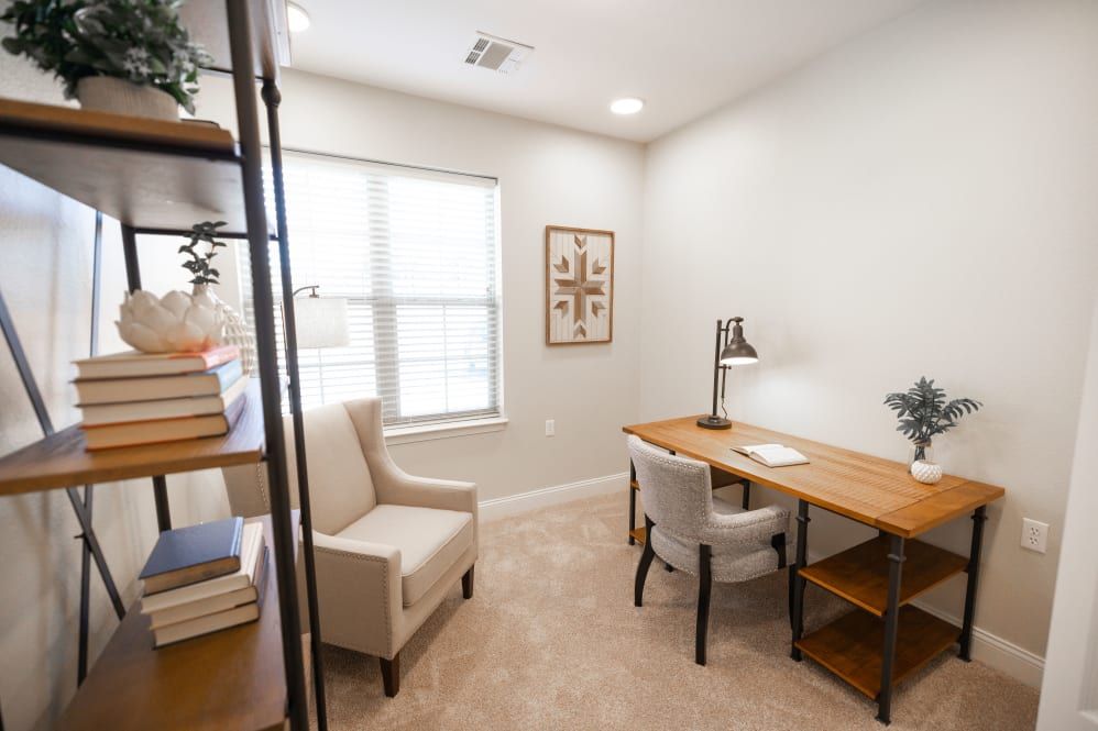 A cozy home office with a wooden desk, upholstered chairs, and a bookshelf by the window.