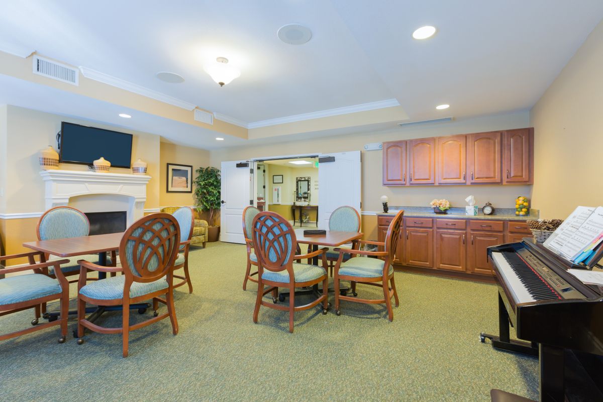 A brightly lit common room with several tables and chairs, featuring a kitchenette area.