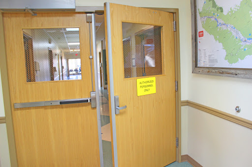 An open wooden door leading to a hallway in a hospital, with a bright yellow sign visible.