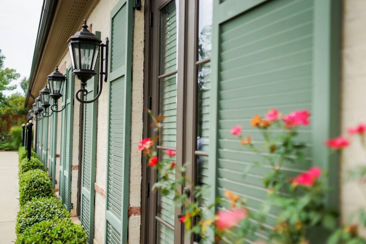 A row of windows with green shutters, adorned by vibrant flowers and a vintage wall lamp.