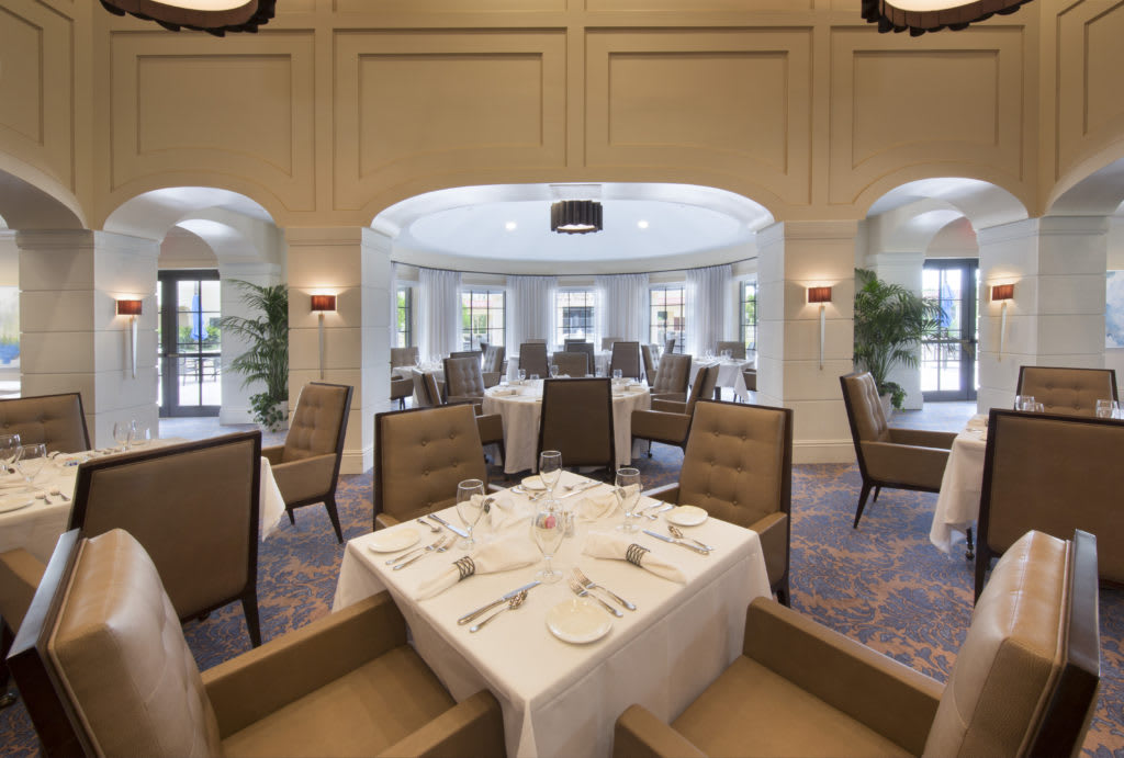 An elegant dining room with neatly set tables and comfortable chairs, featuring large windows that let in natural light.
