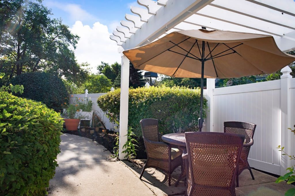 A cozy patio with a shaded seating area under a large umbrella, surrounded by lush greenery on a sunny day.