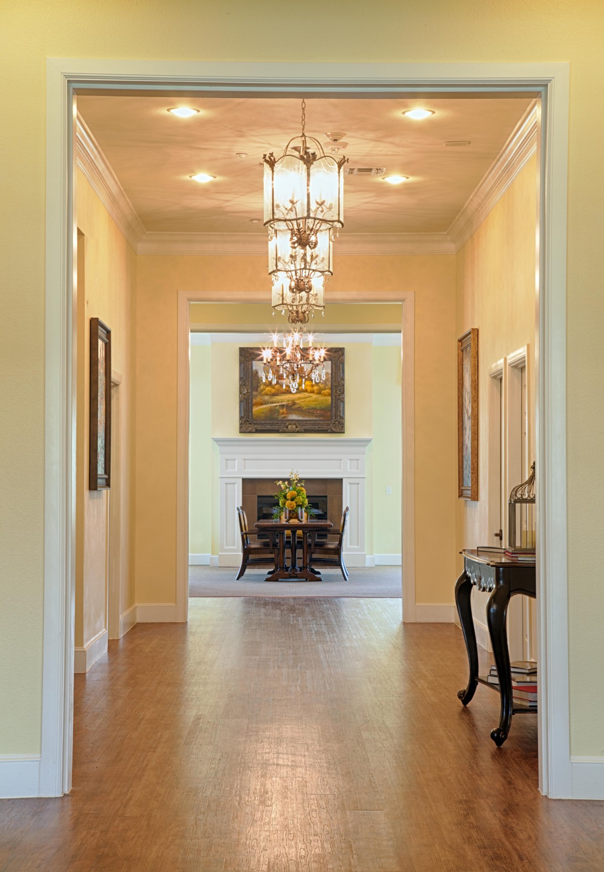 A well-lit hallway with chandeliers, leading to a room featuring a fireplace and artwork.