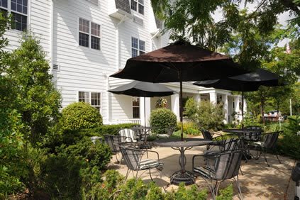 A charming patio area with tables and chairs under large umbrellas, surrounded by lush greenery and adjacent to a white building.