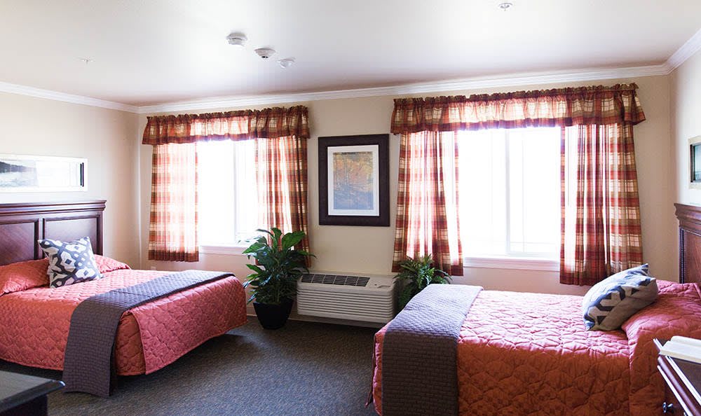A hotel room with two beds, decorated with patterned curtains and a framed painting between large windows.