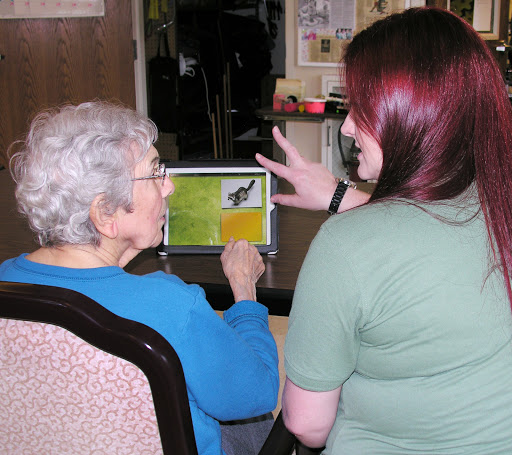 An elderly person engages with a caregiver, exploring content on a tablet together.